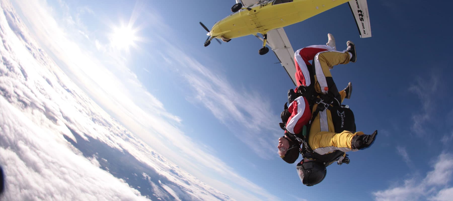 Tandem skydive in freefall after jumping from a yellow airplane above the clouds, with instructor behind the passenger.