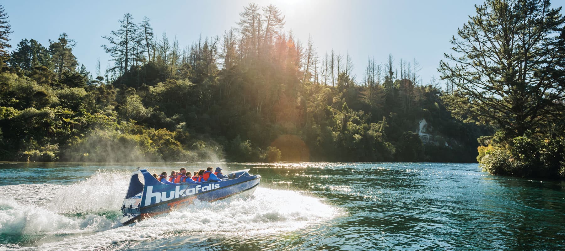  The image shows a blue jet boat labeled "hukafalls" speeding through a calm river, creating a large wake behind it. The boat is filled with passengers wearing life jackets, and it moves swiftly past a scenic landscape of dense green trees and a sunlit fo