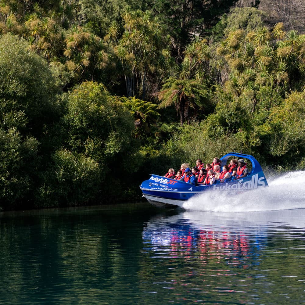 Huka Falls Jet boat with passengers speeding on Waikato River with water spraying behind the boat, surrounded by green trees in Taupō.